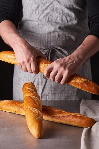 Um homem padeiro com barba em um avental cinza se levanta contra um fundo preto e segura, quebra, corta pão delicioso crocante, pãezinhos, baguete.