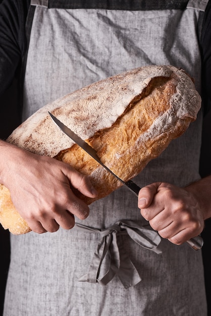 Um homem padeiro com barba em um avental cinza se levanta contra um fundo preto e segura, quebra, corta pão delicioso crocante, pãezinhos, baguete.