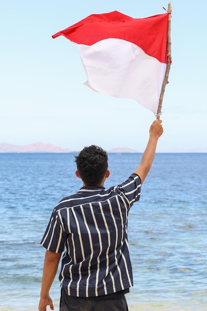 Um homem parado na praia acenando uma bandeira da Indonésia com fundo de paisagem marinha