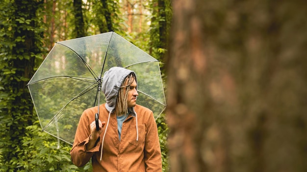 Um homem pensativo com um guarda-chuva na mão na floresta Um hipster com dreadlocks em roupas casuais se esconde da chuva sob um guarda-chuva na floresta