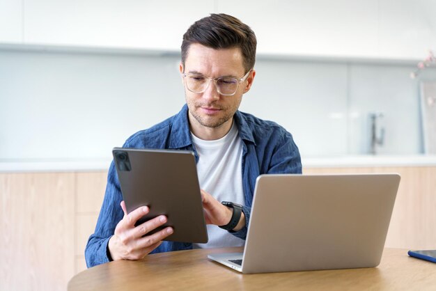 Um homem profissional revisa o conteúdo em um tablet com um laptop aberto diante dele simbolizando
