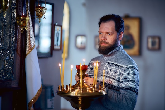 Um homem russo com barba fica em uma Igreja Ortodoxa, acende uma vela e reza na frente do ícone