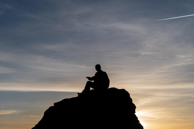 Foto um homem se senta em uma pedra ao pôr do sol, com um pôr do sol ao fundo.