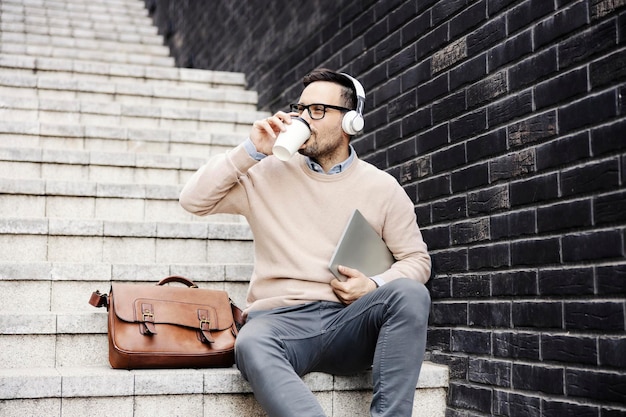 Um homem se senta na escada do lado de fora e bebe café enquanto segura o laptop