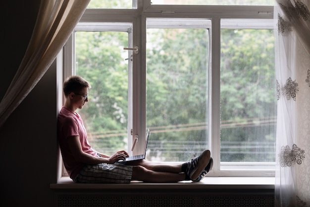 um homem se senta no parapeito de uma janela e trabalha em um laptop