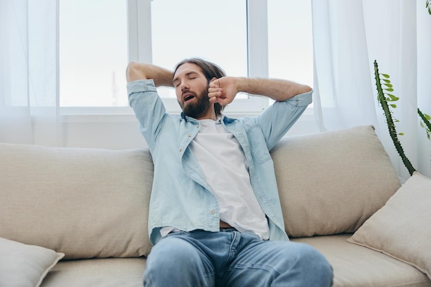 Um homem se senta no sofá bocejando e se espreguiçando após uma soneca, falta de sono e cansaço do trabalho e rotina diária inadequada