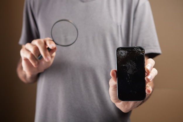 Um homem segurando um telefone quebrado e uma lupa. conceito de pesquisa de conserto de telefone em fundo marrom