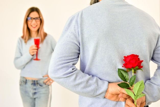 Foto um homem segurando uma rosa vermelha nas costas enquanto sua namorada está de pé no fundo e sorrindo