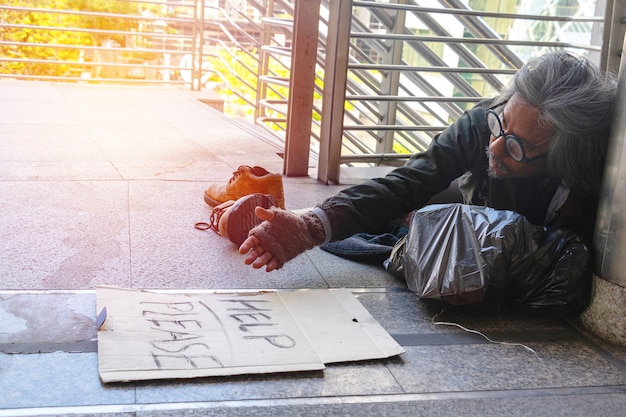 Um homem sem-teto está sentado na passarela na cidade. Ele é mostrar a etiqueta &quot;Ajuda por favor&quot; palavra.