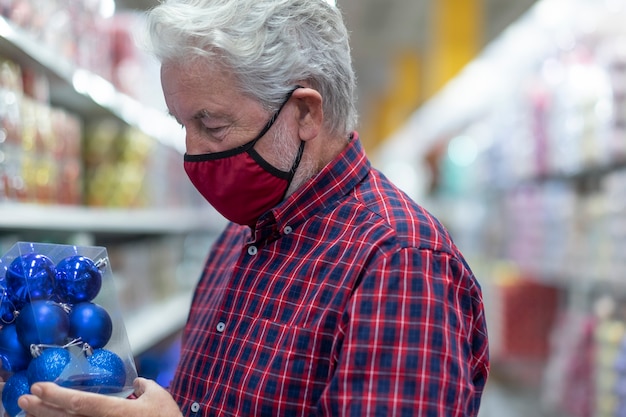 Um homem sênior de cabelos brancos, usando uma máscara médica devido a uma infecção por coronavírus, escolhendo algumas bolas de Natal em uma loja. Novo conceito normal