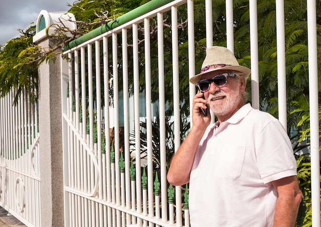 Um homem sênior em pé contra um portão branco no jardim, falando com seu telefone celular. Óculos de sol para o dia de sol.