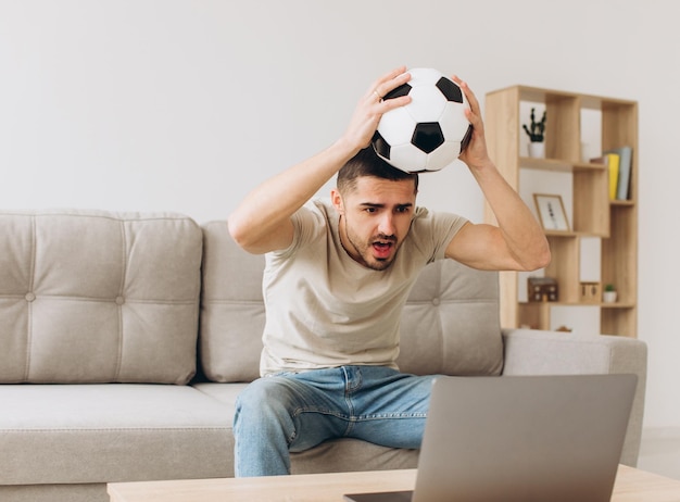 Um homem sentado em um sofá na sala segurando uma bola e torcendo pelo time de futebol assistindo a uma partida em um laptop