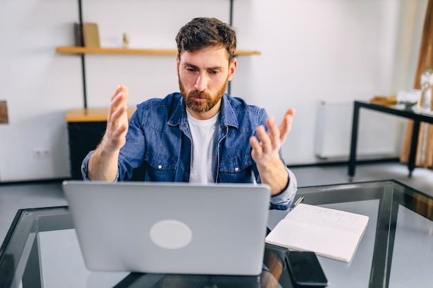 Um homem sentado em uma mesa em casa olhando para um monitor de laptop irritado acenando com as mãos