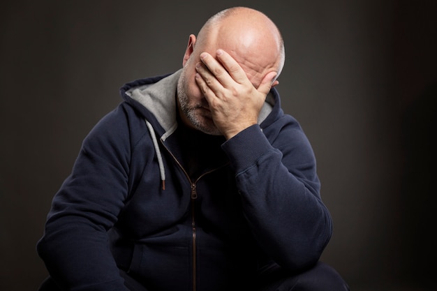 Um homem sério de meia-idade careca, de camiseta preta, está sentado com a mão no rosto. Preto .