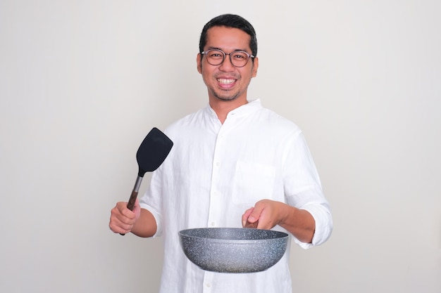 Um homem sorrindo feliz enquanto segura uma frigideira e uma espátula vazias