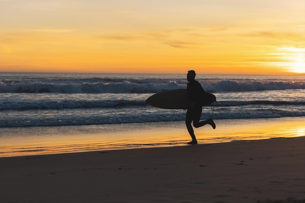 Um homem surfista correndo à beira-mar segurando uma prancha de surf ao pôr do sol