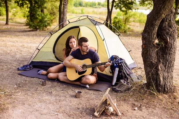 Um homem toca para uma mulher em um violão perto de uma tenda ao pôr do sol. lua de mel na caminhada