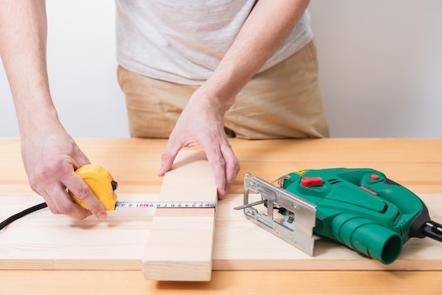 Um homem trabalha com um quebra-cabeça elétrico para madeira em uma mesa de madeira com e sem luvas e mede com uma fita métrica