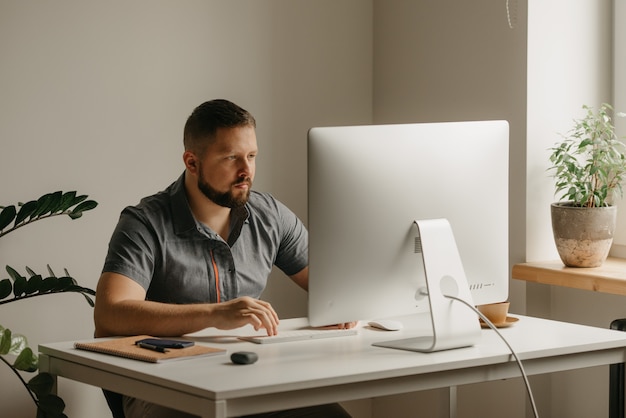 Foto um homem trabalha remotamente em um computador desktop. um cara com barba está digitando durante o relato de um colega em uma videoconferência em casa. um professor está se preparando para uma palestra online.