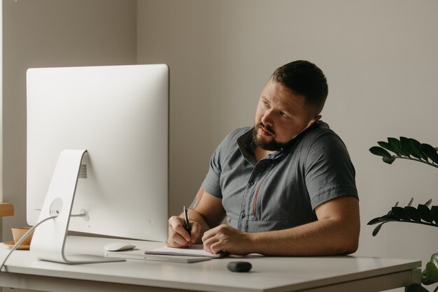 Um homem trabalha remotamente em um computador desktop. um cara de barba está fazendo anotações durante uma ligação telefônica e olhando para a tela do computador em casa. um professor está se preparando para uma palestra online.