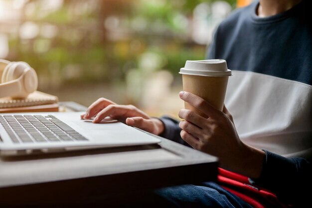 Um homem trabalhando remotamente de uma mesa ao ar livre usando seu laptop enquanto segura uma xícara de café