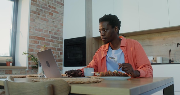 Um homem usa um laptop e almoça em casa.
