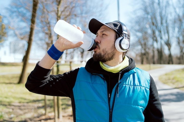 Um homem usando fones de ouvido sem fio bebe água de um atleta de garrafa reutilizável correndo no parque