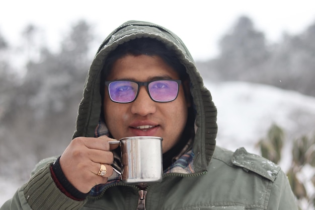 Foto um homem vestindo um moletom verde segura uma xícara de chá em frente a uma paisagem de neve.