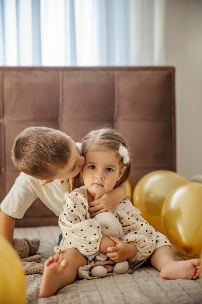 Um irmão está beijando sua irmãzinha enquanto está sentado em uma cama cercado de balões