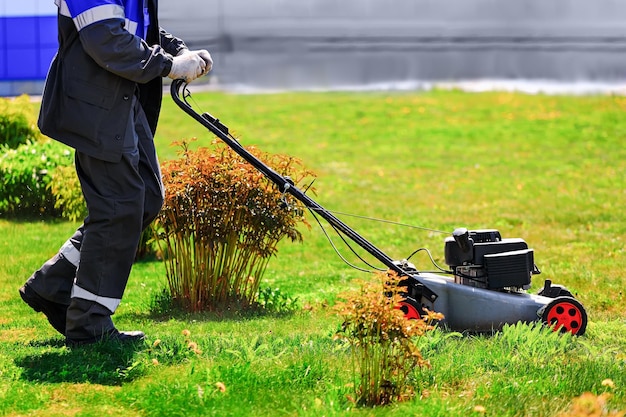 Um jardineiro de macacão caminha com um cortador de grama em um gramado verde um homem corta o gramado em um dia ensolarado de verão