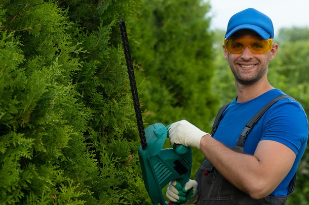 Um jardineiro usando óculos de segurança corta e molda uma cerca viva de thuja com um aparador elétrico