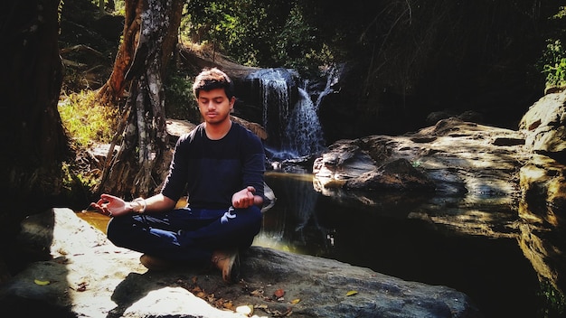 Foto um jovem a meditar numa rocha na floresta.