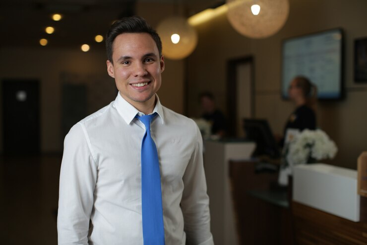 Um jovem administrador de hotel, de camisa branca e gravata azul, fica de pé contra a recepção.