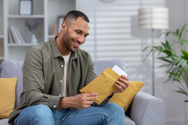 Um jovem africano-americano sorridente está sentado no sofá em casa e abre um envelope no qual ele