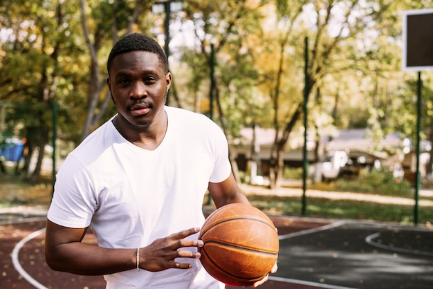 Um jovem afro-americano está jogando basquete em uma quadra ao ar livre, sorrindo e olhando para a câmera. Esportes ao ar livre.