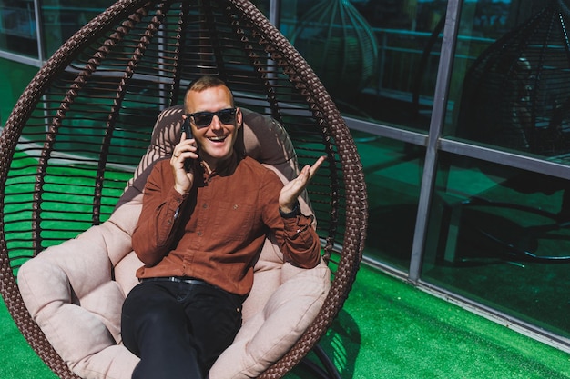 Um jovem alegre de camisa com um telefone senta-se no terraço de um prédio de escritórios depois do trabalho usa internet sem fio um funcionário masculino positivo em óculos está falando ao telefone