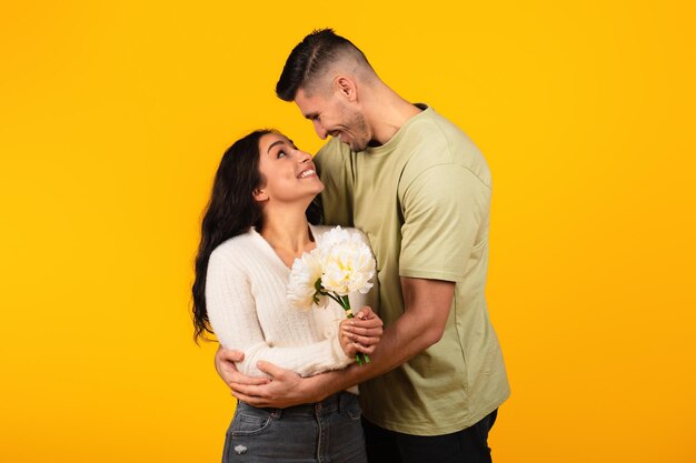 Um jovem árabe alegre em abraços casuais, mulher com flores, desfruta de romântico isolado em fundo amarelo