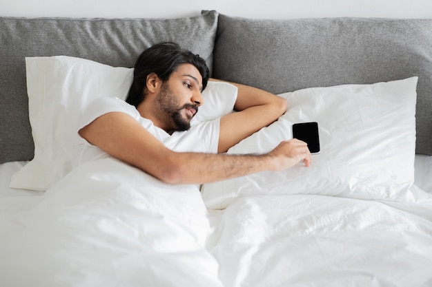 Um jovem árabe sério em camiseta branca dorme acorda deitado na cama usa smartphone com tela em branco