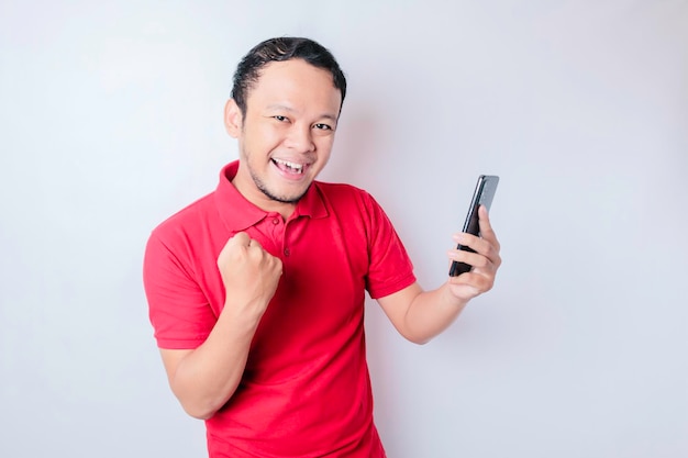 Um jovem asiático com uma expressão feliz e bem-sucedida vestindo camisa vermelha e segurando smartphone isolado por fundo branco