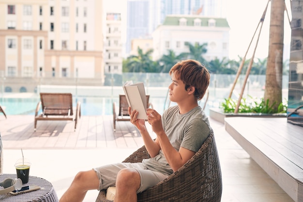 Um jovem asiático está relaxando perto da piscina ao ar livre com um livro nas mãos.