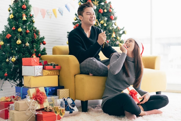 Um jovem asiático feliz soprando um apito de festa nas costas de sua namorada usando um chapéu de Papai Noel segurando um presente recebido em casa com uma árvore de Natal ao fundo