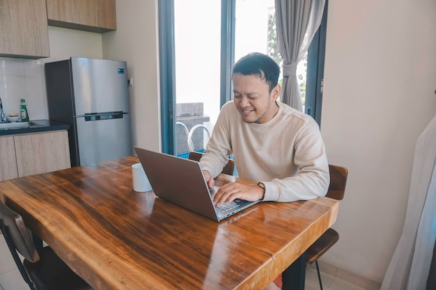 Um jovem asiático passa seu tempo em casa sentado na sala de jantar trabalhando em seu laptop se sente frustrado