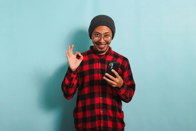 Um jovem asiático satisfeito está usando um smartphone e mostrando um gesto OK isolado em um fundo azul