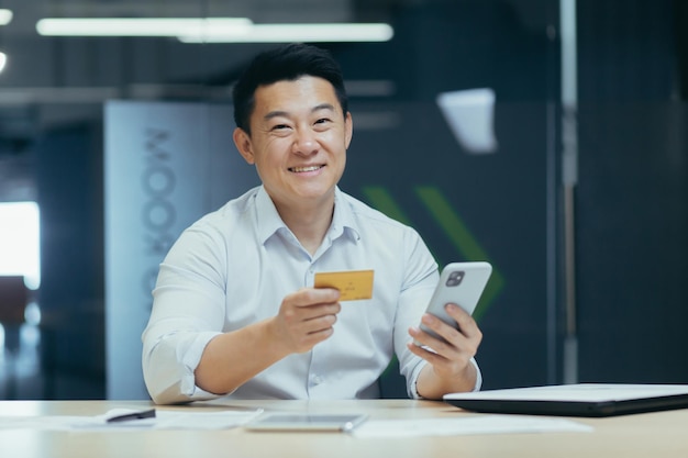 Um jovem asiático trabalha no escritório em uma mesa e segura um cartão de crédito na mão usa o telefone