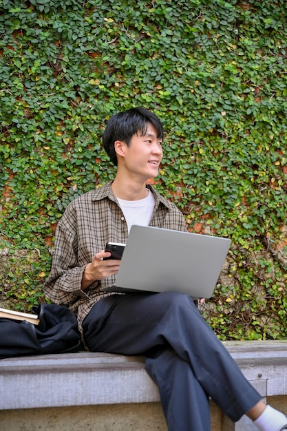 Um jovem asiático trabalhando remotamente no jardim da cidade usando laptop sentado em um banco