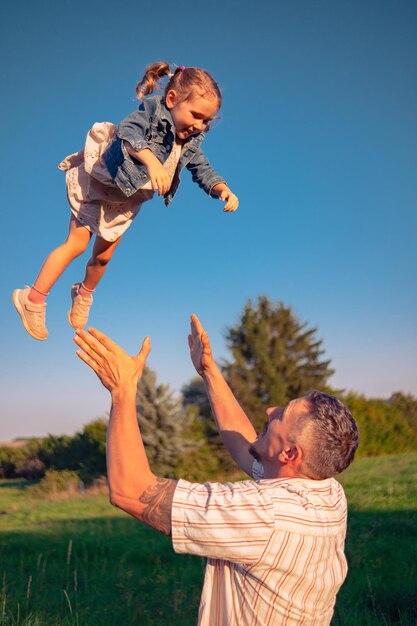 Foto um jovem atira sua filha para cima e ela ri de felicidade pai brincando com sua filha em um prado ensolarado pai e filha passam tempo na natureza