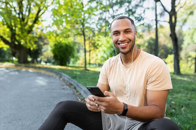Um jovem atleta masculino africano-americano corredor em fones de ouvido senta-se no parque na calçada