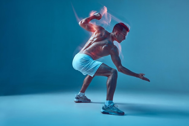 Um jovem atlético musculoso com torso nu executa movimentos de dança no fundo azul do estúdio por muito tempo