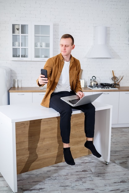 Um jovem atraente com roupas casuais, sentado na cozinha usando um computador laptop. Trabalhe em casa, fluxo de trabalho remoto.