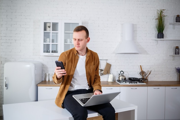 Um jovem atraente com roupas casuais, sentado na cozinha usando um computador laptop. Trabalhe em casa, fluxo de trabalho remoto.